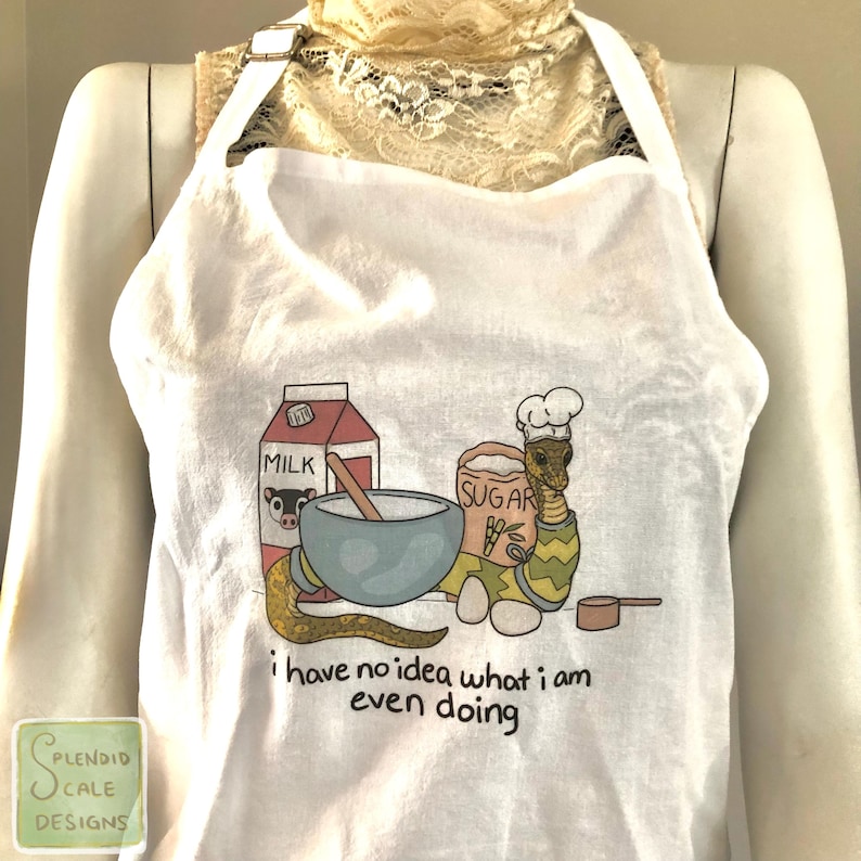 Op de afbeelding: Witte schort met een humoristische illustratie van een slangkok met een mengkom, een melkpak en een suikerzak. De tekst "i have no idea what i am even doing" staat eronder gedrukt. De schort heeft een nekband en strikbanden.