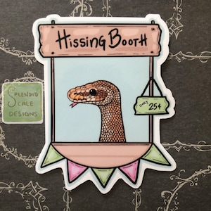 May include: A sticker of a cartoon snake peeking out of a booth with a sign that says "Hissing Booth" and a price tag that says "Only 25¢". The booth is decorated with bunting.