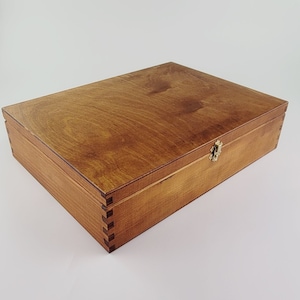 May include: A wooden box with a hinged lid and a brass latch. The box is stained a light brown colour and has a smooth finish.
