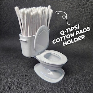 Può includere: Un porta Q-tips e dischetti di cotone grigio a forma di toilette in miniatura. Il sedile del water è aperto e il serbatoio contiene un fascio di bastoncini di cotone bianchi. Il testo "Q-TIPS/ COTTON PADS HOLDER" è scritto in bianco.