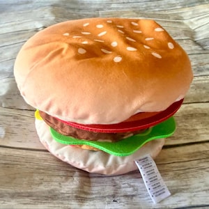 May include: A plush hamburger toy featuring a tan bun with white embroidered sesame seeds, a red felt tomato slice, a brown felt patty, and a green felt lettuce leaf. A white fabric care tag with black text is attached to the bottom bun.
