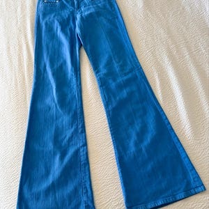 Pode incluir: Jeans flare azul brilhante. Os jeans t&ecirc;m um design cl&aacute;ssico de cinco bolsos, fecho de correr e fecho de bot&atilde;o. O tecido parece ser um denim de peso m&eacute;dio. Os jeans est&atilde;o colocados sobre uma superf&iacute;cie branca.