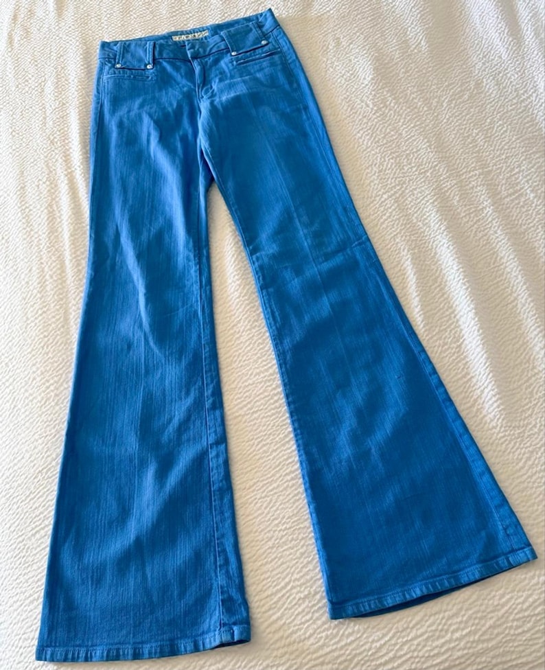 Pode incluir: Jeans flare azul brilhante. Os jeans t&ecirc;m um design cl&aacute;ssico de cinco bolsos, fecho de correr e fecho de bot&atilde;o. O tecido parece ser um denim de peso m&eacute;dio. Os jeans est&atilde;o colocados sobre uma superf&iacute;cie branca.