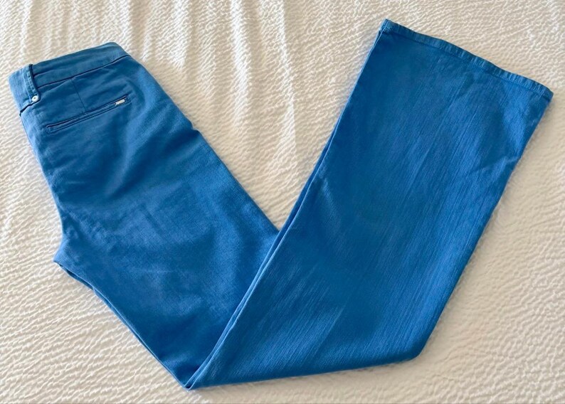 Pode incluir: Cal&ccedil;as azuis claras com design de perna larga. As cal&ccedil;as s&atilde;o feitas de um tecido liso e t&ecirc;m um design cl&aacute;ssico de cinco bolsos. As cal&ccedil;as est&atilde;o colocadas sobre uma superf&iacute;cie branca texturizada.