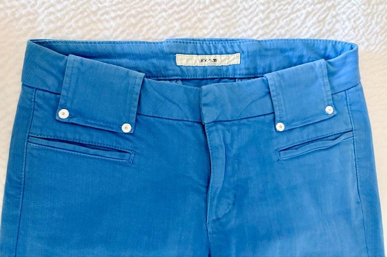 Pode incluir: Close-up de cal&ccedil;as azuis com fecho de bot&atilde;o e dois bolsos frontais. As cal&ccedil;as t&ecirc;m uma cintura sob medida com presilhas e uma pequena etiqueta que diz "JOE'S". As cal&ccedil;as s&atilde;o feitas de um tecido azul liso.