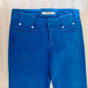 Pode incluir: Cal&ccedil;as azuis brilhantes com corte sob medida. As cal&ccedil;as apresentam um design cl&aacute;ssico com fecho de bot&atilde;o e z&iacute;per, bolsos frontais e uma pequena etiqueta na cintura. A cor vibrante e as linhas limpas tornam estas cal&ccedil;as um item vers&aacute;til.