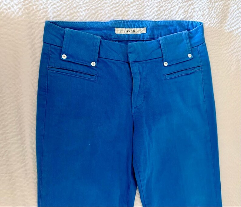 Pode incluir: Cal&ccedil;as azuis brilhantes com corte sob medida. As cal&ccedil;as apresentam um design cl&aacute;ssico com fecho de bot&atilde;o e z&iacute;per, bolsos frontais e uma pequena etiqueta na cintura. A cor vibrante e as linhas limpas tornam estas cal&ccedil;as um item vers&aacute;til.