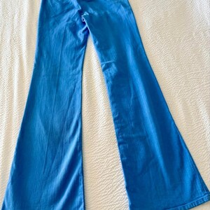 Pode incluir: Cal&ccedil;as boca de sino azul claro. As cal&ccedil;as s&atilde;o feitas de um material semelhante ao denim e t&ecirc;m um design cl&aacute;ssico de cinco bolsos. As cal&ccedil;as est&atilde;o colocadas sobre uma superf&iacute;cie branca, mostrando sua forma e detalhes.