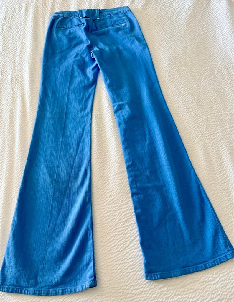 Pode incluir: Cal&ccedil;as boca de sino azul claro. As cal&ccedil;as s&atilde;o feitas de um material semelhante ao denim e t&ecirc;m um design cl&aacute;ssico de cinco bolsos. As cal&ccedil;as est&atilde;o colocadas sobre uma superf&iacute;cie branca, mostrando sua forma e detalhes.