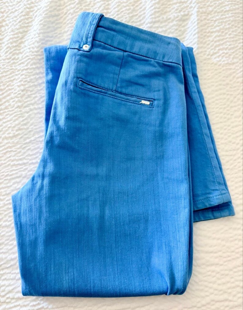 Pode incluir: Um par de cal&ccedil;as azuis claras dobradas. As cal&ccedil;as t&ecirc;m fecho de bot&atilde;o, um pequeno bolso nas costas e uma pequena etiqueta com o log&oacute;tipo. As cal&ccedil;as s&atilde;o feitas de um tecido texturizado e est&atilde;o cuidadosamente dobradas sobre uma superf&iacute;cie branca.