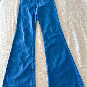 Pode incluir: Jeans flare azul brilhante. Os jeans t&ecirc;m um design cl&aacute;ssico de cinco bolsos com fecho de bot&atilde;o e z&iacute;per. O tecido parece ser um denim de peso m&eacute;dio. Os jeans est&atilde;o colocados sobre uma superf&iacute;cie branca.