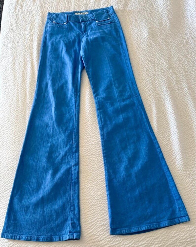 Pode incluir: Jeans flare azul brilhante. Os jeans t&ecirc;m um design cl&aacute;ssico de cinco bolsos com fecho de bot&atilde;o e z&iacute;per. O tecido parece ser um denim de peso m&eacute;dio. Os jeans est&atilde;o colocados sobre uma superf&iacute;cie branca.