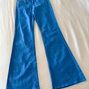 Pode incluir: Cal&ccedil;as de ganga azul brilhante com pernas largas. As cal&ccedil;as t&ecirc;m um design cl&aacute;ssico de cinco bolsos, fecho de bot&atilde;o e z&iacute;per e perna larga. O tecido parece ser ganga de peso m&eacute;dio. As cal&ccedil;as est&atilde;o estendidas numa superf&iacute;cie branca.