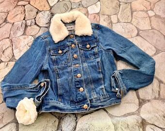 May include: A blue denim jacket with a cream-colored faux fur collar. The jacket has gold-colored buttons and pockets. The sleeves are cuffed, and a white flower is placed near the cuff. The jacket is a classic style.