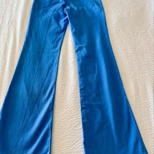 Pode incluir: Cal&ccedil;as boca de sino azul brilhante. As cal&ccedil;as t&ecirc;m cintura alta e abertura larga na perna. A parte de tr&aacute;s das cal&ccedil;as tem dois bolsos e uma presilha para cinto. O tecido tem um ligeiro brilho. As cal&ccedil;as est&atilde;o estendidas numa superf&iacute;cie branca.