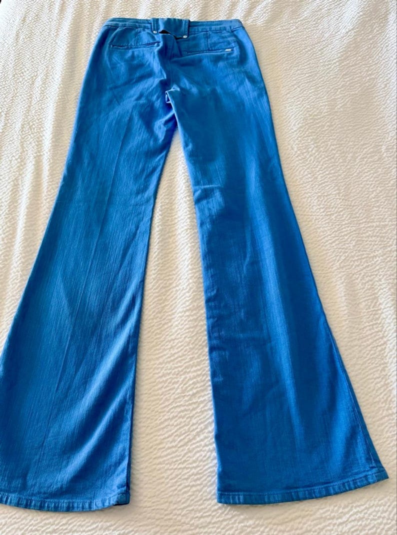 Pode incluir: Cal&ccedil;as boca de sino azul brilhante. As cal&ccedil;as t&ecirc;m cintura alta e abertura larga na perna. A parte de tr&aacute;s das cal&ccedil;as tem dois bolsos e uma presilha para cinto. O tecido tem um ligeiro brilho. As cal&ccedil;as est&atilde;o estendidas numa superf&iacute;cie branca.