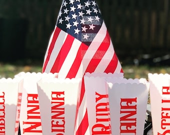 May include: A row of white popcorn containers with red scalloped edges. Each container has a name written in red letters. The names are "Finley", "Nina", "Sienna", "Ridy", "Liena", and "Stella". An American flag is behind the containers.