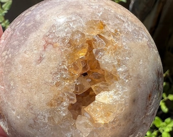 Puede incluir: Una gran geoda de ágata redonda y transparente con una gran abertura que revela un cúmulo de cristales de cuarzo más pequeños y transparentes en su interior. La geoda es de color marrón claro con toques de rosa y blanco.