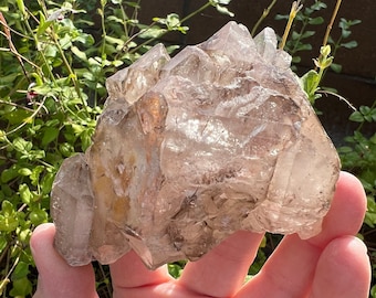 May include: A large, clear quartz crystal cluster with a light brown hue. The crystal has many points and facets, and it is illuminated by the sun.