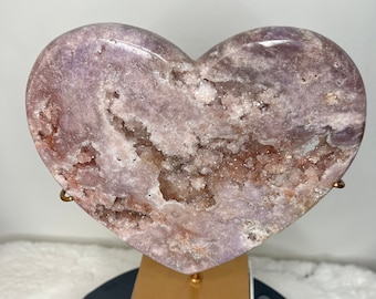 May include: A heart-shaped amethyst crystal with a natural druzy finish, displayed on a gold stand. The crystal is a light purple color with white and pink accents.