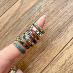 Könnte beinhalten: Eine Sammlung bunter Emaille-Ringe mit goldfarbenen Bändern. Die Ringe weisen verschiedene Blumen- und abstrakte Muster in Blau-, Grün-, Rot-, Weiß- und Schwarztönen auf. Die Ringe werden auf einem Finger vor einem Holzgrund gezeigt.