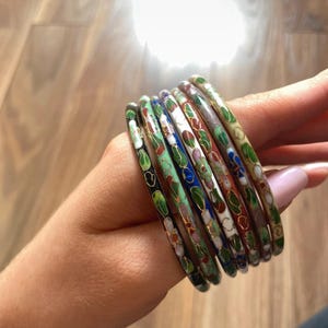 Könnte beinhalten: Eine Sammlung von sieben bunten Emaille-Armreifen. Jeder Armreif zeigt ein Blumenmuster mit verschiedenen Farben wie Grün, Blau, Rot und Weiß. Die Armreifen werden auf einer Hand präsentiert.