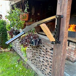 May include: A robust, black and silver log splitter attached to a wooden post, designed for splitting firewood. The background features a wooden structure, plants, and a fire pit with flames visible.