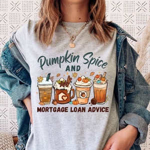 May include: A heather gray t-shirt with the text "Pumpkin Spice and Mortgage Loan Advice" above an illustration of four different pumpkin spice lattes. The design includes fall leaves and pumpkins. The shirt is worn with a denim jacket.