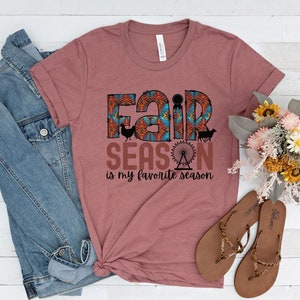 May include: A mauve t-shirt with a graphic print that says "Fair Season is my favorite season". The graphic features a colorful fairground design with a Ferris wheel, a chicken, a pig, and a goat.