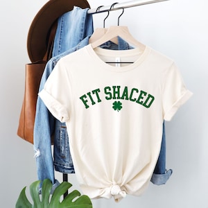 May include: Cream-colored t-shirt with the words "FIT SHACED" in green, arched above a green shamrock. The shirt is hanging on a wooden hanger. A denim jacket and a brown hat are visible in the background.