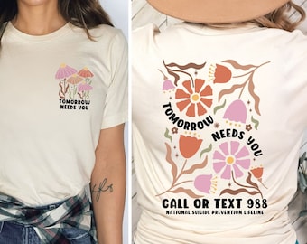 May include: A cream-colored t-shirt with a floral design and the text "Tomorrow Needs You" on the front. The back of the shirt has a larger floral design with the same text and the text "Call or Text 988 National Suicide Prevention Lifeline".