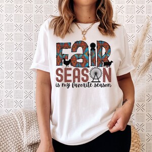 May include: White t-shirt with a colorful graphic that says "Fair Season is my favorite season". The graphic includes a Ferris wheel, a chicken, and a cow.