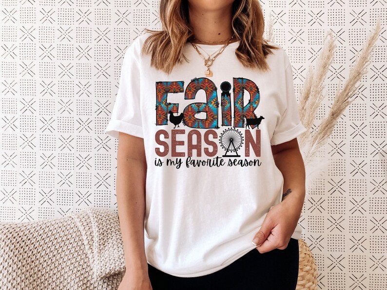 May include: White t-shirt with a colorful graphic that says "Fair Season is my favorite season". The graphic includes a Ferris wheel, a chicken, and a cow.