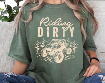 Może przedstawiać: Zielony T-shirt z napisem "Riding Dirty" w stylu vintage. Pod napisem znajduje się kreskówkowy obrazek samochodu terenowego.