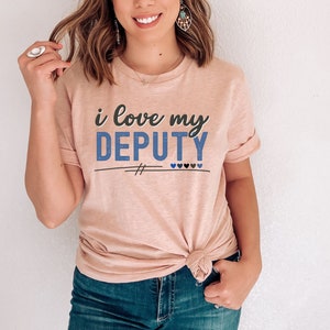 Pode incluir: Uma camiseta rosa com o texto "i love my DEPUTY" em letras azuis e pretas. O texto &eacute; sublinhado com duas linhas pretas e tem quatro pequenos cora&ccedil;&otilde;es azuis abaixo.