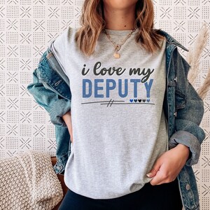 Pode incluir: Uma camiseta cinza com o texto "i love my DEPUTY" em letras azuis e pretas. H&aacute; quatro pequenos cora&ccedil;&otilde;es azuis abaixo do texto.