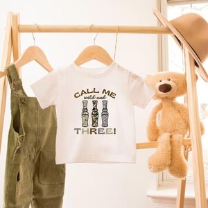 Puede incluir: Camiseta blanca con un estampado marr&oacute;n, negro y blanco que dice "Call me wild and three!" con tres botellas estilizadas. La camiseta est&aacute; colgada en un perchero de madera.