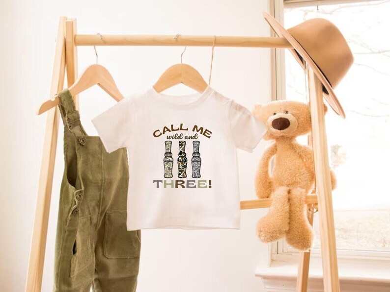 Puede incluir: Camiseta blanca con un estampado marr&oacute;n, negro y blanco que dice "Call me wild and three!" con tres botellas estilizadas. La camiseta est&aacute; colgada en un perchero de madera.