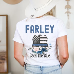 Pode incluir: Camiseta branca com o texto "FARLEY" e um distintivo de pol&iacute;cia azul e branco com uma linha azul fina e detalhes florais. O texto "Back The Blue" est&aacute; abaixo do distintivo.