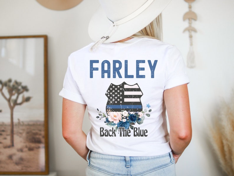 Pode incluir: Camiseta branca com o texto "FARLEY" e um distintivo de pol&iacute;cia azul e branco com uma linha azul fina e detalhes florais. O texto "Back The Blue" est&aacute; abaixo do distintivo.