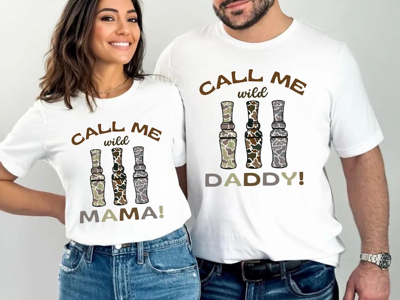 Puede incluir: Dos camisetas blancas con un estampado de camuflaje marr&oacute;n y verde de llamadas de pato. El texto en las camisetas dice "Call me wild" y "Daddy!" o "Mama!"