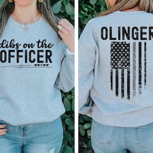 May include: A gray sweatshirt with the text "dibs on the OFFICER" on the front and a thin black line with three hearts underneath. The back of the sweatshirt has the text "OLINGER" above a distressed American flag with a thin gray stripe down the middle.