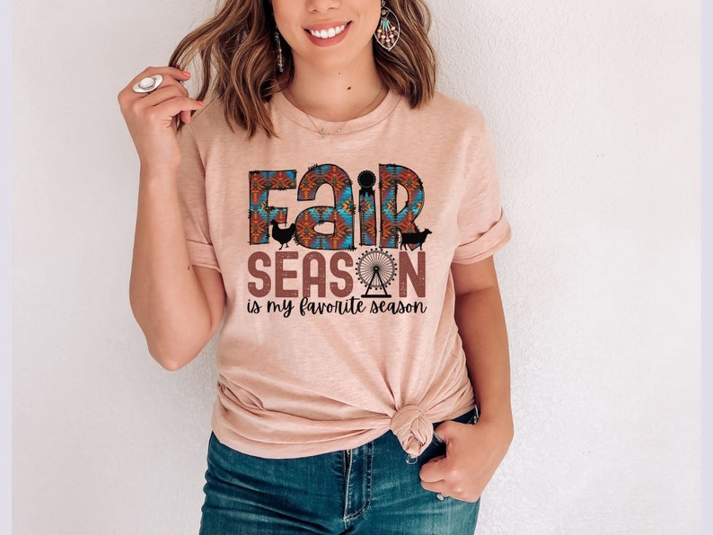 May include: A light pink t-shirt with a graphic design that says "Fair Season is my favorite season". The graphic features a colorful pattern with a Ferris wheel, a chicken, and a sheep.