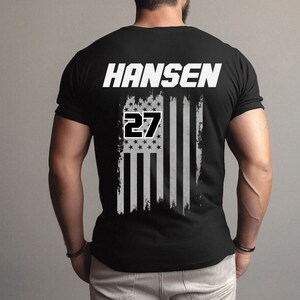 Może przedstawiać: Czarny T-shirt z nadrukiem białej, przetartej flagi amerykańskiej i tekstem "HANSEN 27" na plecach.