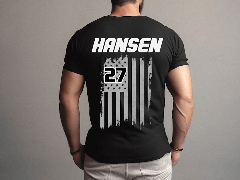 Może przedstawiać: Czarny T-shirt z nadrukiem białej, przetartej flagi amerykańskiej i tekstem "HANSEN 27" na plecach.