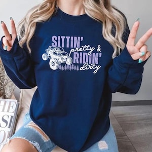 May include: Navy blue sweatshirt with the text "Sittin' pretty & ridin' dirty" in white and purple. The graphic features a white off-road vehicle and a row of trees. The sweatshirt is a casual style.