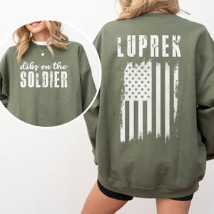 May include: A green sweatshirt with a white distressed American flag and the text "LUPREK" on the back. The front of the sweatshirt has the text "dibs on the SOLDIER" in white.