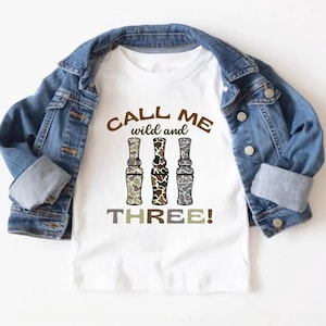Puede incluir: Camiseta blanca con un estampado de camuflaje marr&oacute;n, blanco y gris de tres llamadas de pato. El texto "Call me wild and three!" est&aacute; impreso en la camiseta.