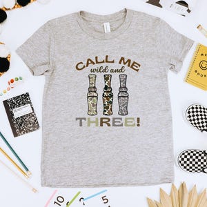Puede incluir: Una camiseta gris con el texto "Call me wild and three!" impreso en ella. El texto est&aacute; rodeado de tres im&aacute;genes de llamadas de pato de camuflaje.