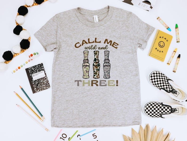 Puede incluir: Una camiseta gris con el texto "Call me wild and three!" impreso en ella. El texto est&aacute; rodeado de tres im&aacute;genes de llamadas de pato de camuflaje.